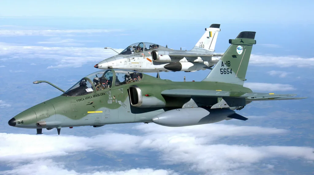 Due caccia bombardieri biposto Embraer A-1B AMX della Força Aérea Brasileira in volo in formazione serrata sopra le nuvole, uno con mimetica verde-grigia e uno grigio chiaro.