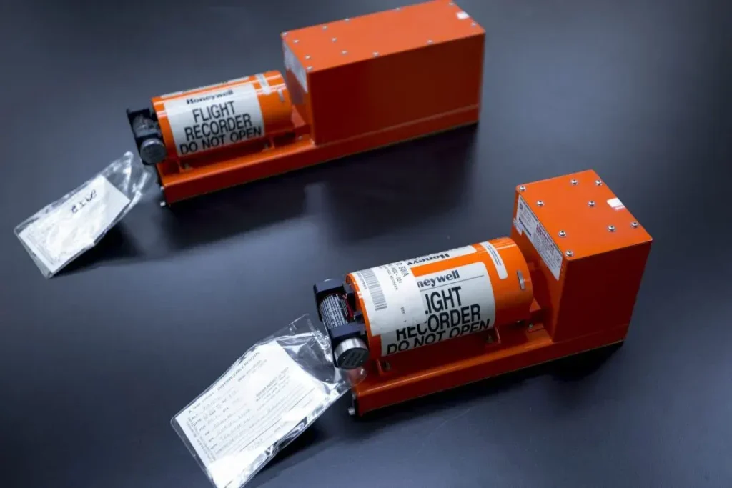 Due scatole nere aeronautiche Honeywell arancioni su un tavolo nero. Primo piano sui cilindri di memoria con la scritta "FLIGHT RECORDER DO NOT OPEN" e sui localizzatori acustici subacquei metallici neri e argento fissati all'estremità.