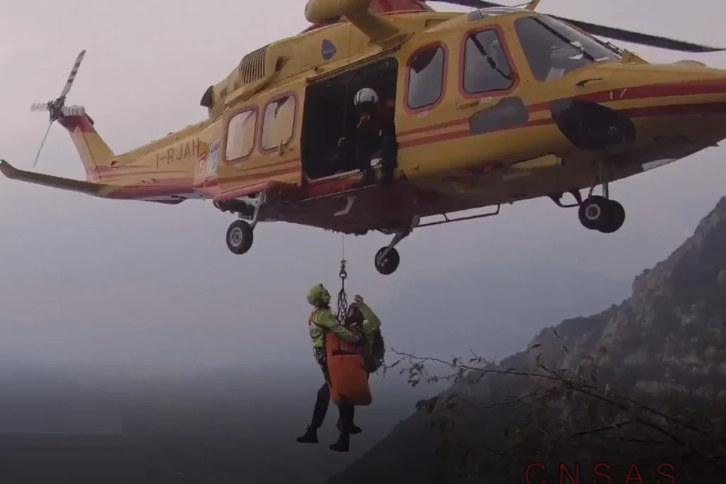 Un elicottero da soccorso bimotore Leonardo AW139 (marche I-RJAH), gestito da Avincis Aviation Italia e impiegato nei servizi di elisoccorso per conto del 118 della Regione Lombardia (base di Brescia). L'immagine illustra una complessa operazione di recupero in ambiente impervio tramite verricello, condotta in sinergia con i tecnici del CNSAS (Corpo Nazionale Soccorso Alpino e Speleologico).